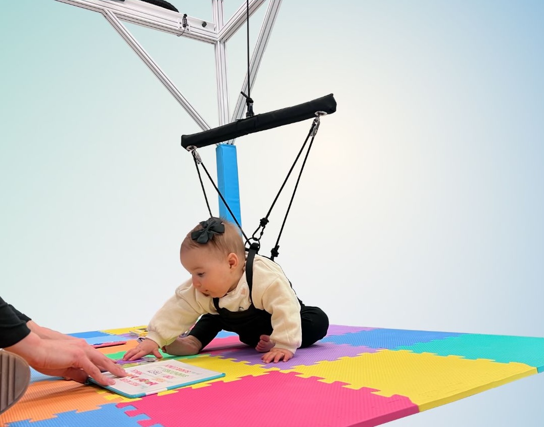 An infant in a BabyG harness reaching for a book on a play mat