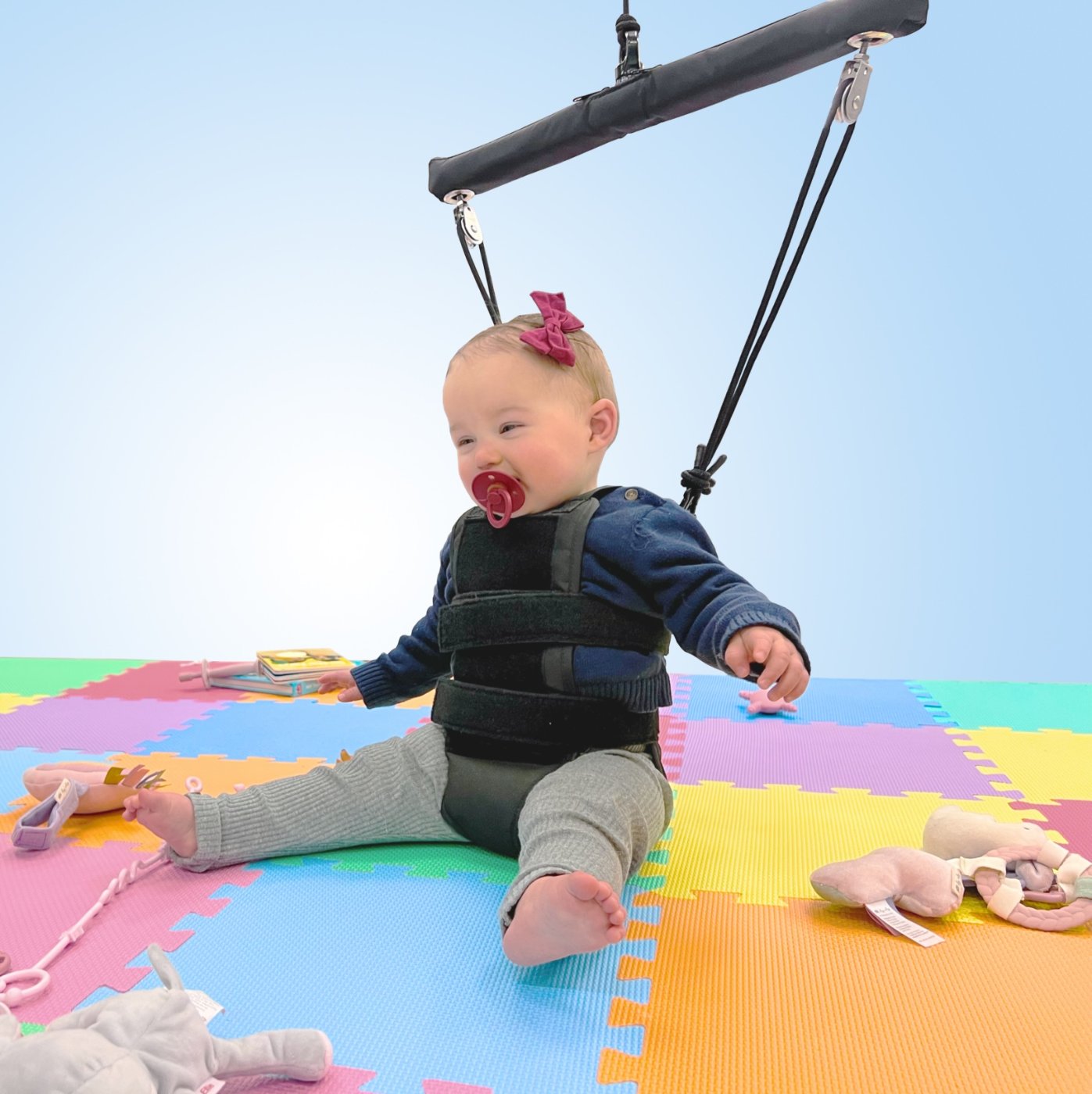 An infant seated on a play mat wearing a BabyG harness suspended from the overhead system